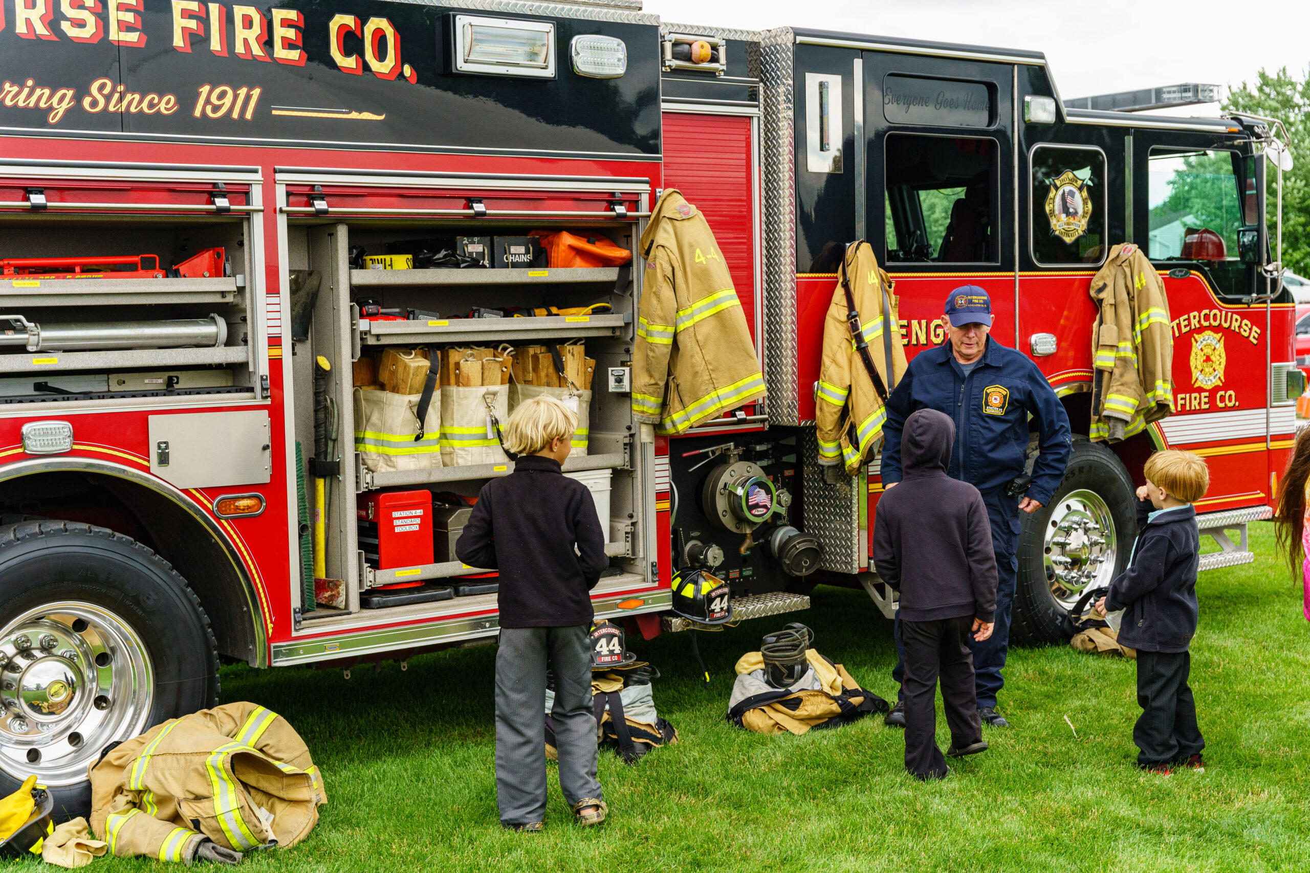 Firefighter speaking with three boys in front of a fire engine with all the compartments open.